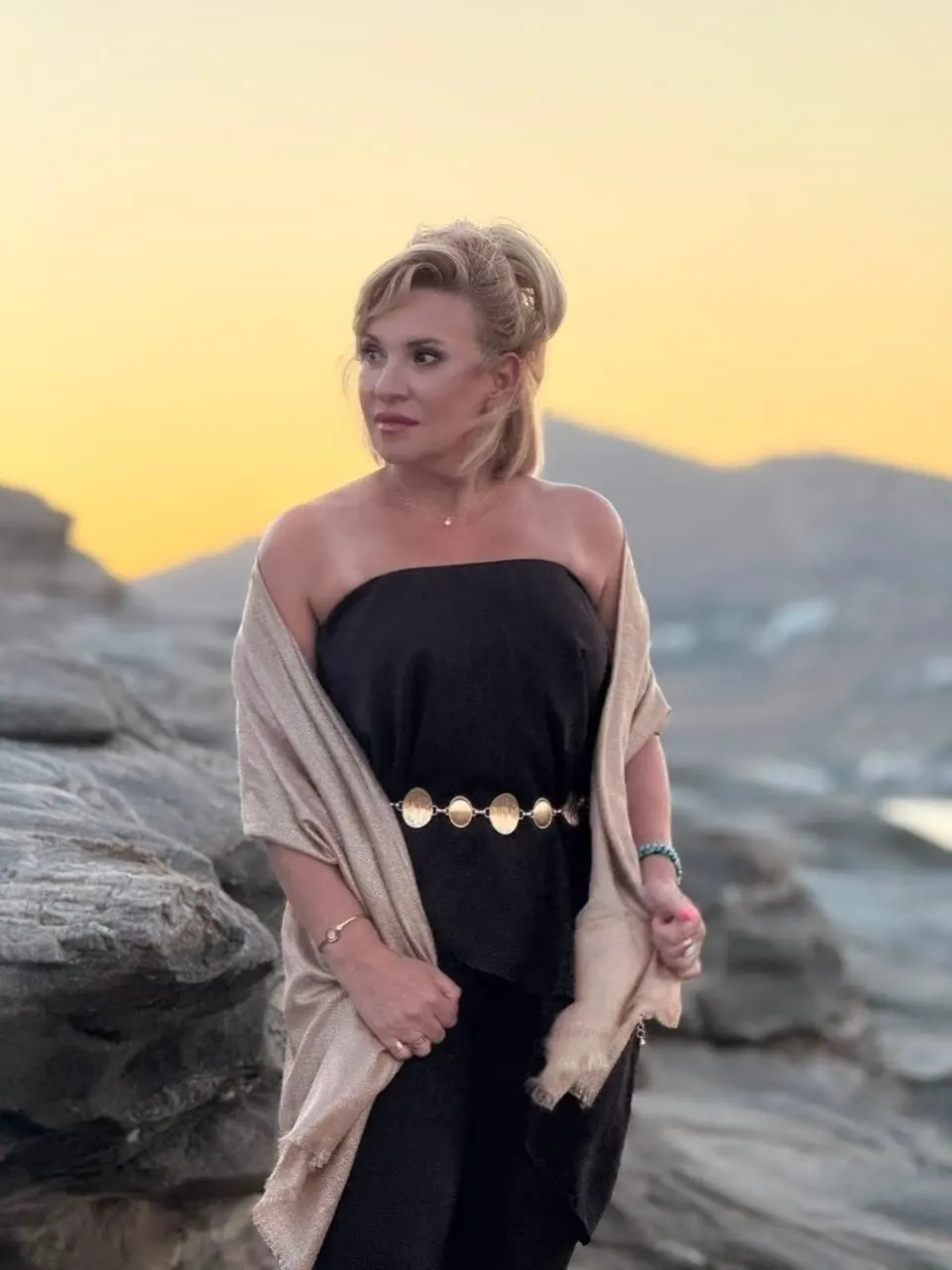 Amalia Kyrou standing on coastal rocks at sunset, a light shawl over a dark top, looking out to one side
