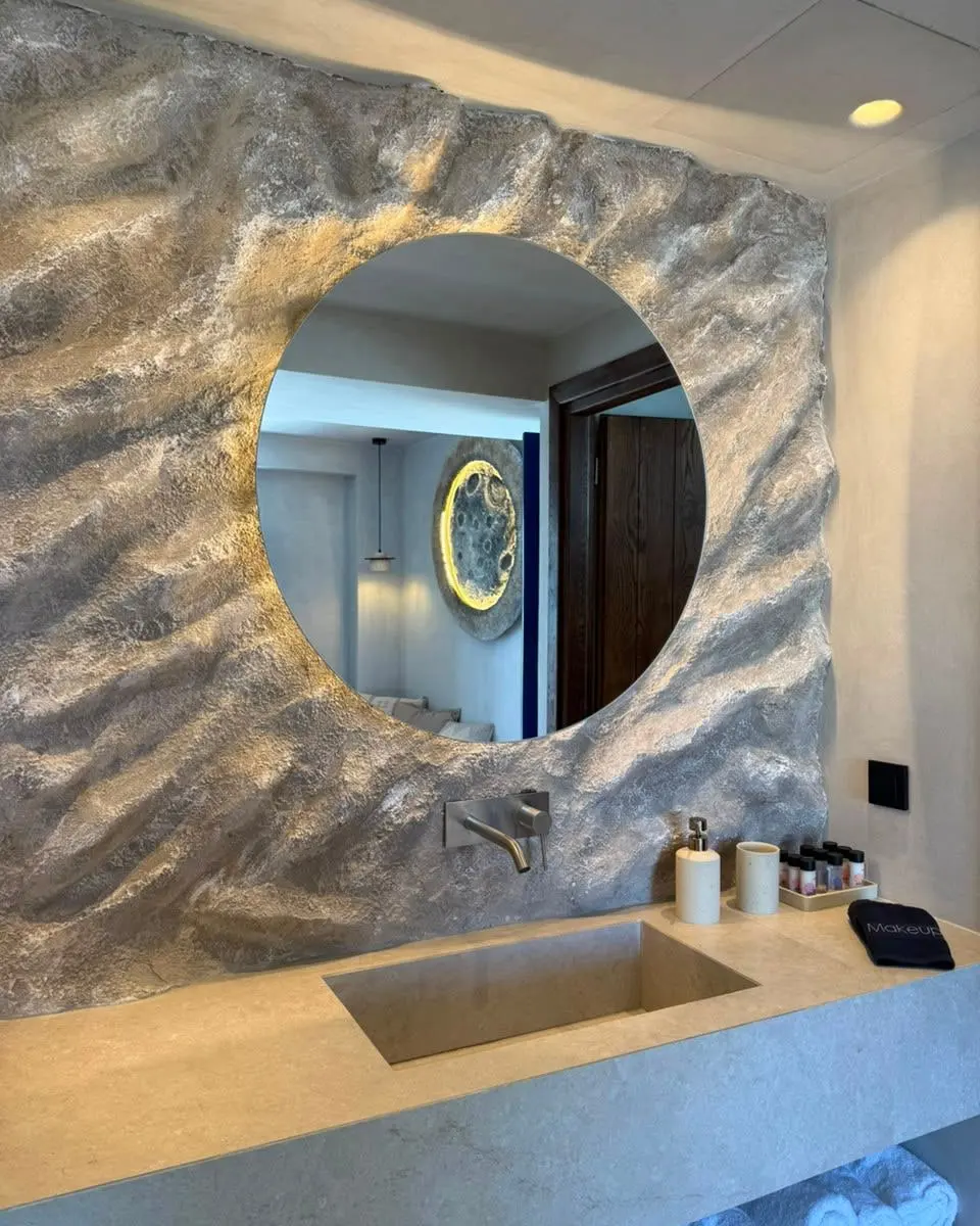 A round backlit mirror set into the sculpted rock wall above a stone basin in the bathroom