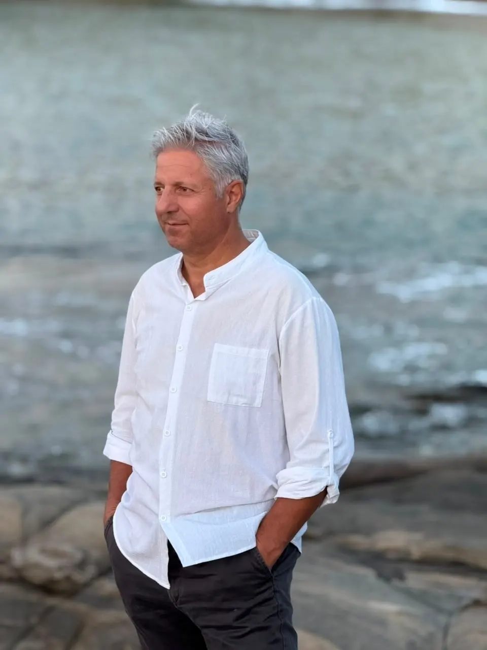 Panagiotis Savvidis in a white linen shirt, hands in his pockets, standing on flat rocks by the sea